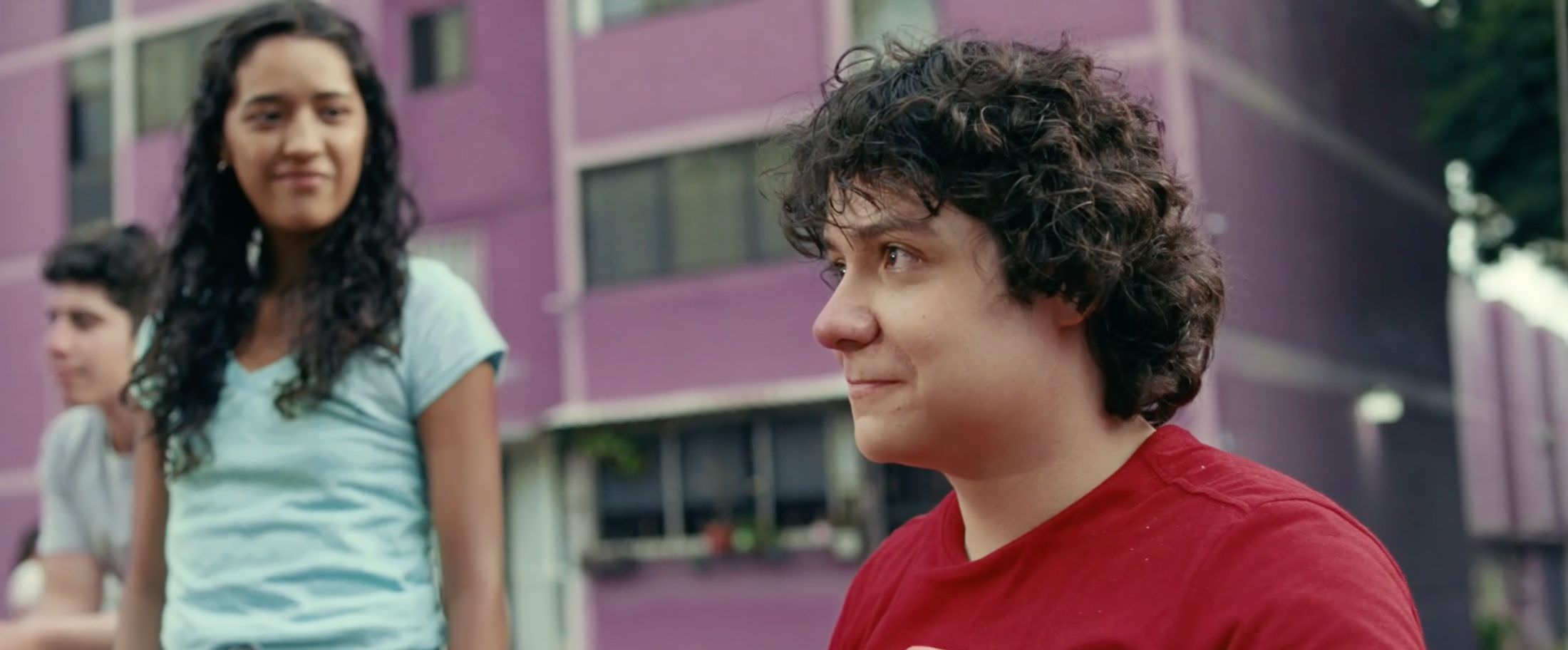 A young man (off-screen, he is in a wheelchair) looks forward with determination as his friends look proudly at him in the background